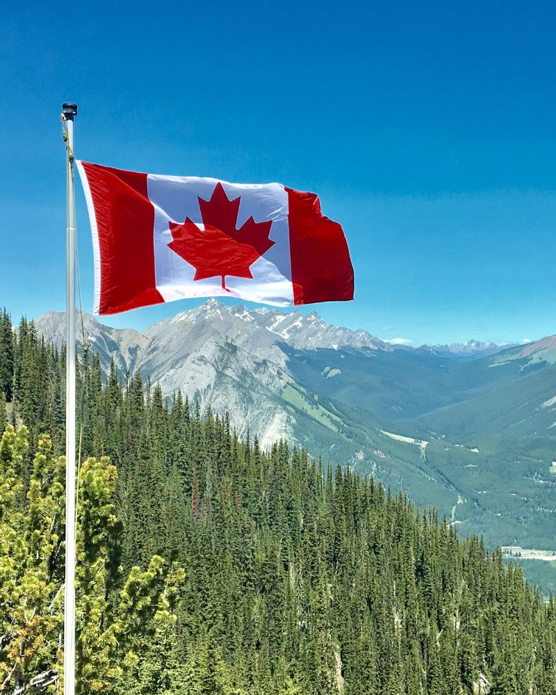 A Canadian flag flutters on a flagpole with a backdrop of a forested mountainous landscape under a clear blue sky, symbolizing the welcoming spirit of Canada and its robust Provincial Nominee Programs for immigration.