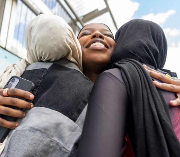 Three people are hugging; two wearing hijabs, one smiling in the middle. One person is holding a smartphone. They are outside under a glass canopy with a blue sky in the background, celebrating their recently acquired citizenship status.