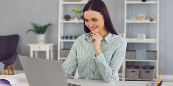 A woman in a striped shirt sits at her desk, looking at a laptop and smiling. Perhaps she's exploring opportunities with the International Mobility Program. Behind her, office shelves and a plant add to the professional yet inviting atmosphere.