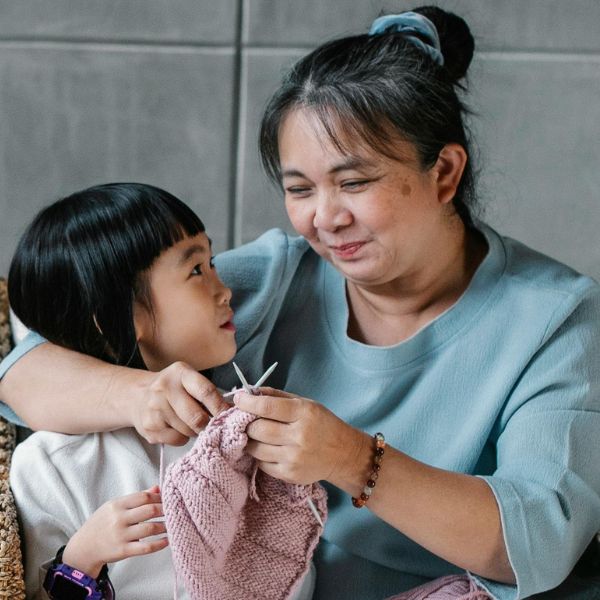 An older woman and a young girl sit together, smiling, as they knit a pink fabric piece indoors, celebrating the warmth of family sponsorship.