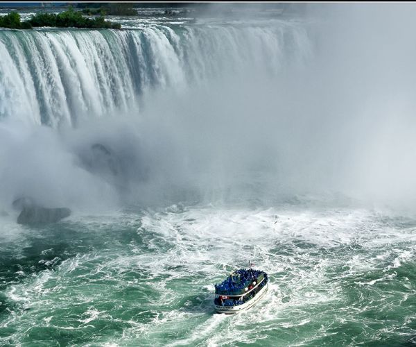 A boat with international tourists approaches the base of a large waterfall in Canada, surrounded by mist and churning water, offering an unforgettable experience.