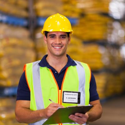 A worker in a high-visibility vest and hard hat smiles while holding a clipboard, feeling at home as if he just gained permanent residence in this bustling warehouse.