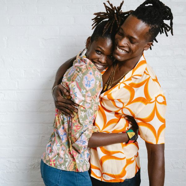 A smiling couple in patterned shirts embraces warmly in front of a white brick wall, celebrating their new Family Visa.