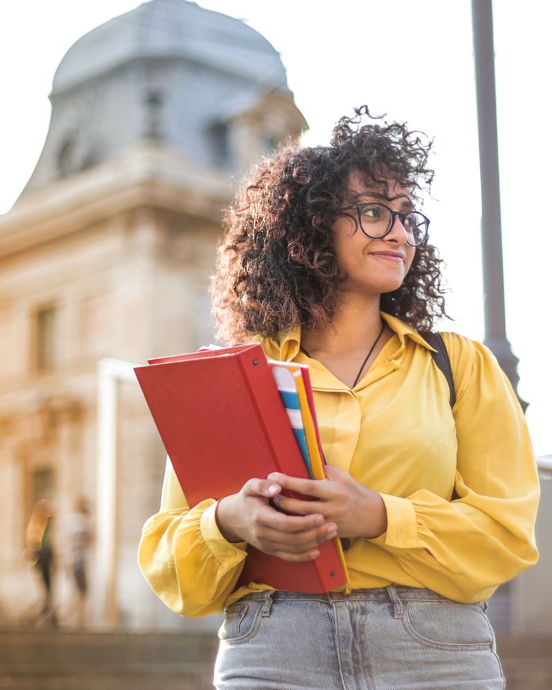 A person with curly hair and glasses, wearing a yellow shirt, stands outside holding folders. A historic building is in the background, adding an international flair reminiscent of Canada's rich heritage.