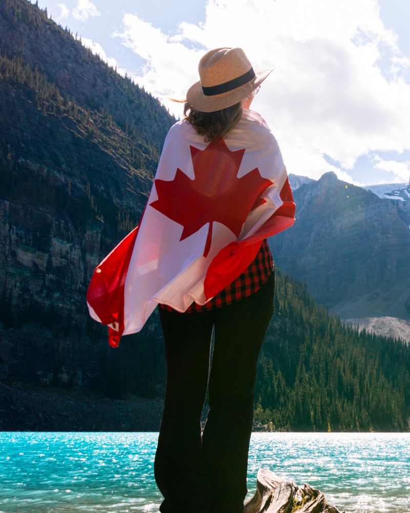 A traveler in a straw hat and red plaid shirt stands by a tranquil blue lake, proudly draped in a Canadian flag. The stunning mountain and forest backdrop capture the spirit of Canada, inviting all who embrace adventure and tourism to explore its natural wonders.