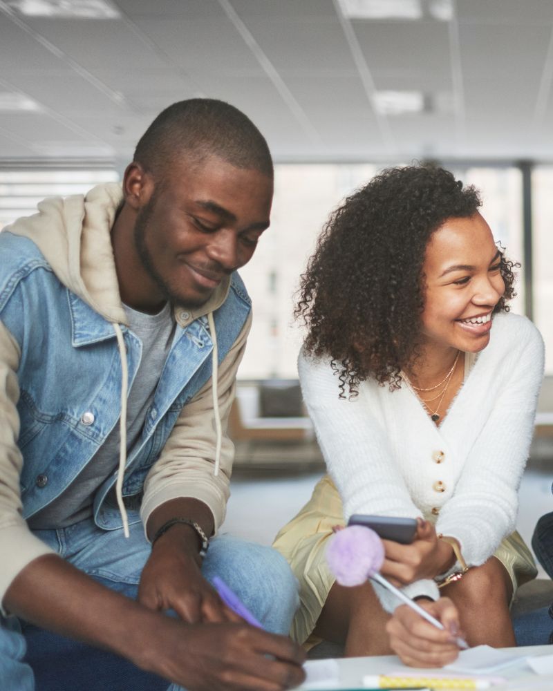 Two people are smiling and working together at a table, one writing while the other, granted open work permits, holds a phone.