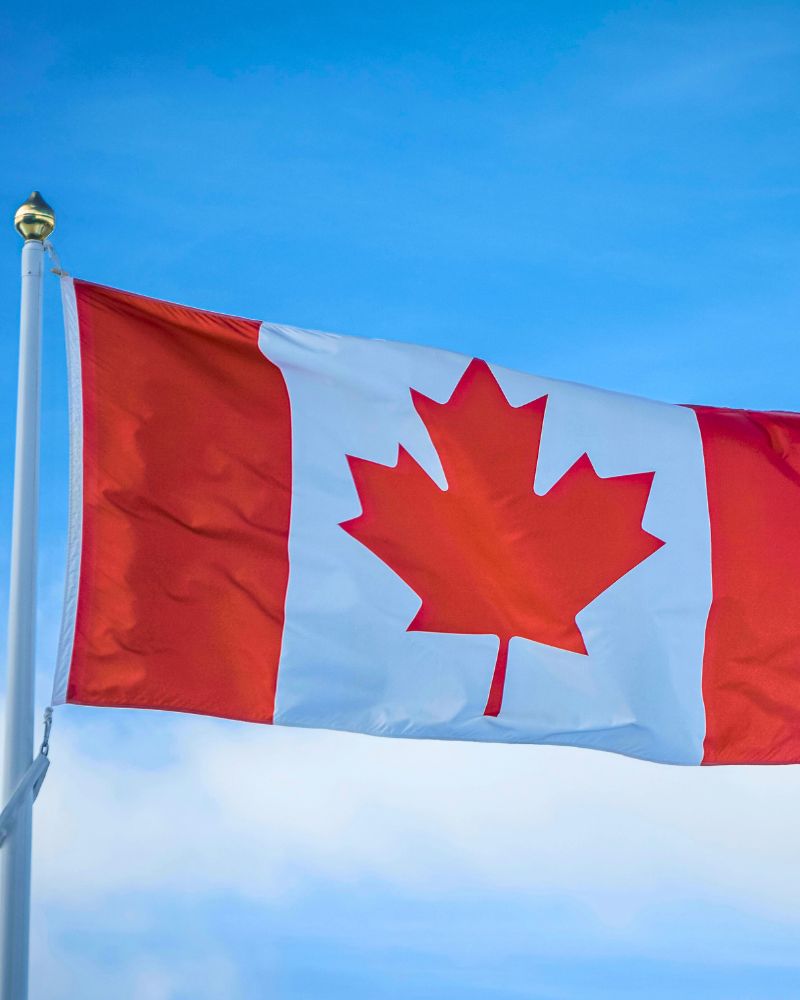 The Canadian flag, with its iconic red maple leaf flanked by two red vertical stripes, waves proudly against a clear blue sky. It's a symbol of opportunity for many pursuing dreams through avenues like open work permits.