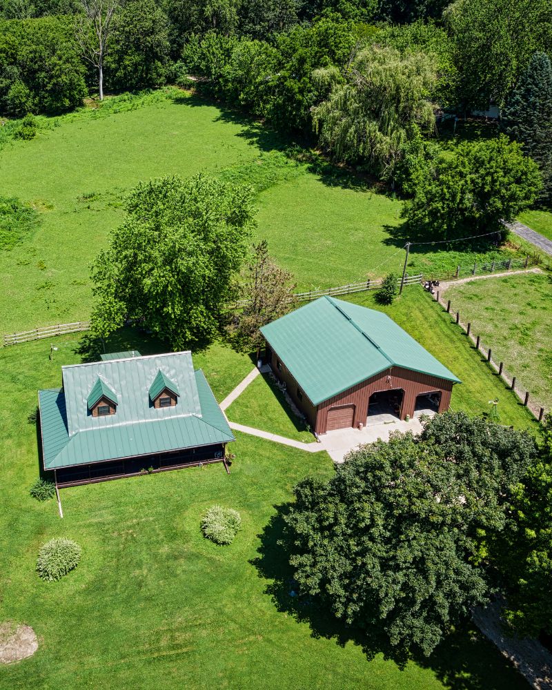 Aerial view of a rural property with two green-roofed buildings surrounded by trees and fields, embodying the peaceful landscapes that many seek through programs like the Saskatchewan Immigrant Nominee Program.