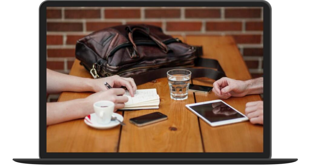 Two people at a wooden table with a notebook, smartphone, tablet, glass of water, coffee cup, and bag.