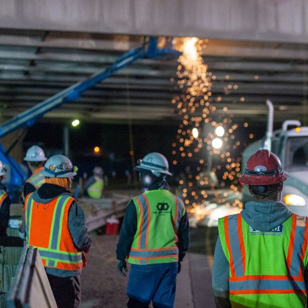 Construction workers in safety vests and helmets stand under a bridge at night as sparks fly from welding, demonstrating the necessity of skilled labor in projects that often require careful assessment like an LMIA.